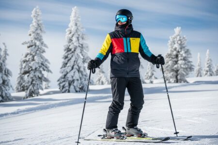 Osoba v zimnej výstroji stojí na zasneženej zjazdovke s lyžami a palicami za jasného dňa.